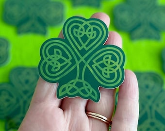 May include: A hand holding a green shamrock sticker with a Celtic knot design. The sticker is surrounded by other shamrock stickers on a green background.