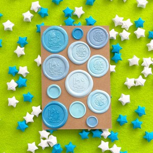 May include: A rectangular brown card featuring various circular wax seals in shades of blue. Some seals display the text Eid Mubarak with a mosque silhouette, while others feature intricate Arabic calligraphy. Small paper stars surround the card.