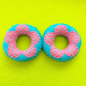 Può includere: Due ciambelle di feltro fatte a mano. Ogni ciambella ha una base turchese con glassa rosa a forma di fiore e zuccherini blu. Le ciambelle sono ambientate su uno sfondo verde-giallo brillante, creando una composizione giocosa e colorata.