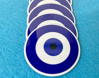 May include: A stack of blue and white evil eye stickers. The stickers are round and have a white center with a blue eye in the middle. The stickers are arranged in a vertical stack.