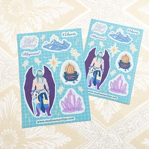 May include: Two sticker sheets with a light blue grid pattern. Each sheet features illustrations of a winged figure, mountains, stars, crystals, and a cauldron. Text includes "Hello Fae Darling", "Rhysand", and "Polaris". The website address www.studioamarillys.com is also visible.