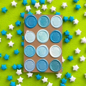 May include: A rectangular brown card featuring twelve circular wax seals arranged in four rows of three. Each seal displays a raised Hamsa hand symbol in varying shades of blue, ranging from light sky blue to deep navy, with a textured finish.
