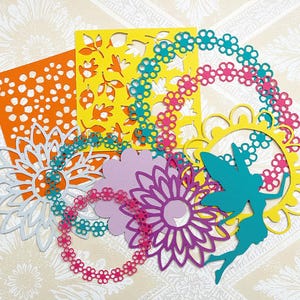 May include: A collection of colorful paper cutouts in various shapes. Included are orange, yellow, purple, and white floral designs, along with a teal fairy silhouette and flower chain accents. The cutouts are arranged on a patterned background.