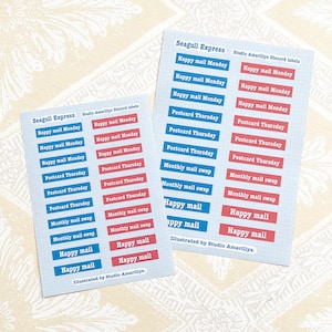 Può includere: Due fogli di adesivi con etichette rettangolari rosse e blu. Le etichette mostrano frasi come "Happy mail Monday" e "Postcard Thursday". È visibile anche il testo "Seagull Express" e "Illustrated by Studio Amarillys".