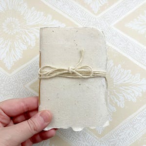 May include: A small, handmade notebook with a rough, textured, cream-colored paper cover. The notebook is tied closed with a piece of twine.