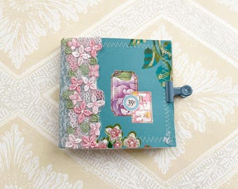 Può includere: Un piccolo diario quadrato con una copertina turchese ornata da illustrazioni floreali e pizzo decorativo. Il dorso presenta pizzo floreale rosa e bianco. Una clip blu fissa il diario. La copertina include francobolli in stile vintage.
