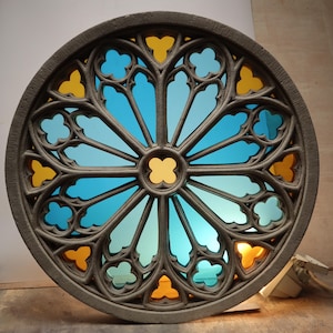 May include: A circular, gray stone window with a stained glass design. The glass features a blue and yellow color scheme, with a central yellow cross-shaped design. The window has a detailed, gothic-style frame.