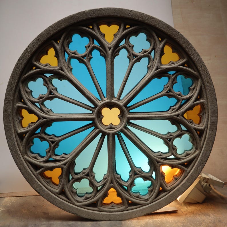 May include: A circular, gray stone window with a stained glass design. The glass features a blue and yellow color scheme, with a central yellow cross-shaped design. The window has a detailed, gothic-style frame.