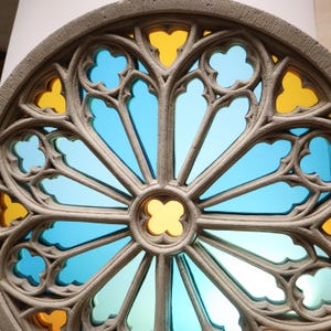 May include: A circular stained glass window with a stone frame. The window features a symmetrical design with blue and yellow glass panes. The stone frame has a textured surface and a detailed, ornate pattern.