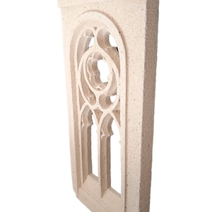 May include: A white stone window frame with a decorative, intricate design. The frame is made of a rough, textured material and has a slightly arched top.