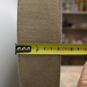 May include: A close-up of a beige, textured rectangular object being measured with a yellow tape measure. The tape measure shows a measurement of approximately 3 inches.
