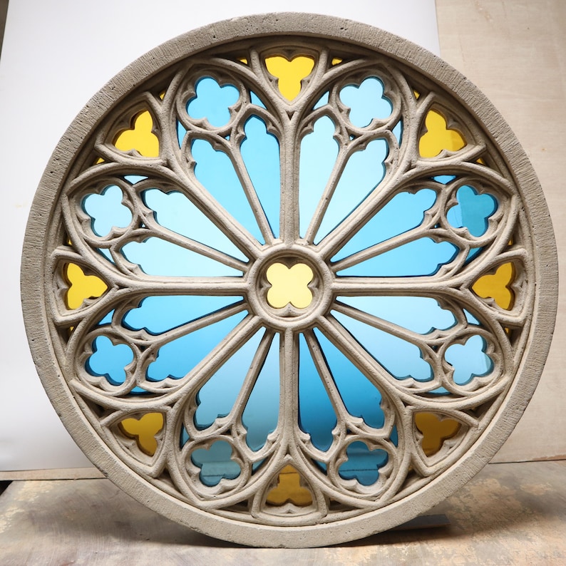 May include: Circular stone window with a stained glass design. The window features a light gray frame with a repeating pattern of blue and yellow glass panes. The center of the window has a yellow quatrefoil design.