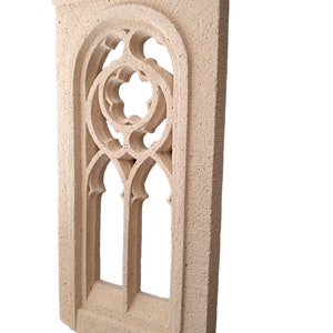 May include: A beige stone wall decor with a carved window design. The window has a pointed arch and a decorative pattern of circles and leaves.