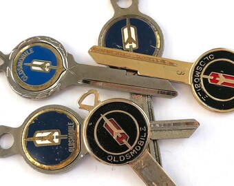 May include: Five vintage Oldsmobile car keys with different colored logos. The keys are silver and gold with black, red, and blue accents. The logos feature the words "Oldsmobile" and "Classic".