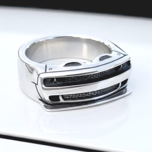 May include: A silver-colored ring with a car grill design. The ring has a wide band and a detailed front resembling a car's bumper and headlights. The ring is displayed on a white surface.