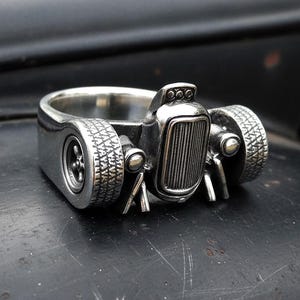 Puede incluir: Un anillo plateado con forma de coche hot rod clásico. El anillo presenta neumáticos en miniatura detallados, una parrilla delantera y otros componentes del coche. El anillo está sobre un fondo oscuro, resaltando su acabado metálico y diseño.