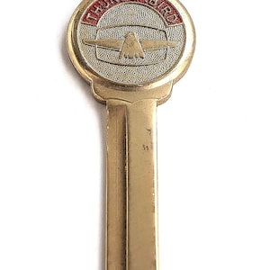 May include: A brass-colored metal key featuring a circular head with a textured surface. The head displays a stylized bird emblem and the word THUNDERBIRD in red lettering. The key has a standard blade with a central groove and a small loop at the top.