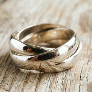 May include: A silver-colored ring with three interlocking bands. The bands are smooth and reflective, creating a bright, shiny appearance. The ring is set against a rustic, wooden surface.