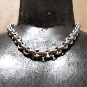May include: A silver-colored chain necklace with a series of interlocking oval links. The necklace is displayed on a dark, textured surface, showcasing its metallic sheen and design. The chain is a classic jewelry piece.