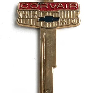 Könnte beinhalten: Ein goldfarbener Metallschlüssel mit einem dekorativen Kopf, der den Schriftzug CORVAIR in roten Buchstaben trägt. Unter dem Text befindet sich ein blaues Chevrolet-Emblem auf einem strukturierten goldenen Hintergrund mit vertikalen Linien.