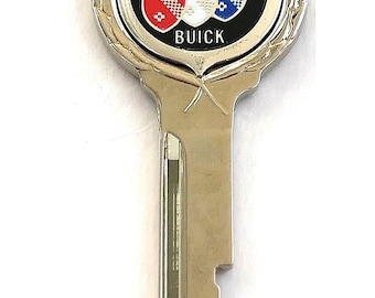 Può includere: Una chiave in metallo argentato con una testa circolare e un inserto nero. Il centro mostra il logo Buick con tre scudi in rosso, bianco e blu, e la parola BUICK stampata in lettere bianche sotto gli scudi su uno sfondo nero.