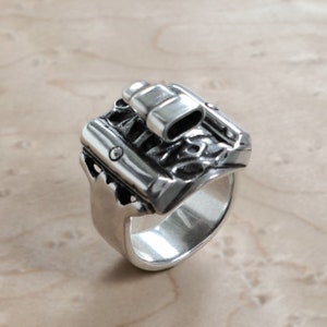 Puede incluir: Un anillo plateado con un diseño detallado y texturizado. El anillo presenta una parte superior rectangular con elementos cilíndricos y ovalados, y una banda ancha. El anillo está hecho de un material metálico brillante.