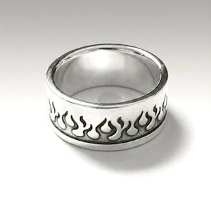 Könnte beinhalten: Ein silberner Ring mit Flammenmuster. Der Ring hat ein breites Band mit polierter Oberfläche. Das Flammenmuster ist um den Umfang herum geätzt und bietet eine auffällige Ästhetik. Geeignet für den täglichen Gebrauch, ist der Ring ein mutiges Statement-Stück.