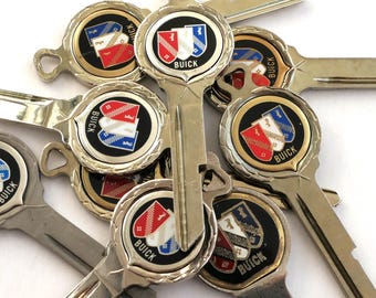 May include: A collection of vintage Buick car keys. Each key features a circular emblem with the Buick logo and a shield design in red, white, and blue. The keys are silver-toned and have a classic design.