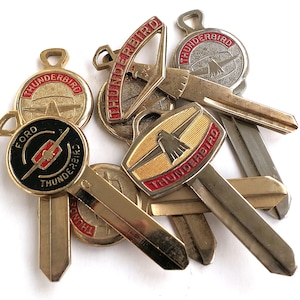 May include: A collection of six vintage gold-toned keys with red and black accents. The keys feature the words "Thunderbird" and "Ford" on the key tops.