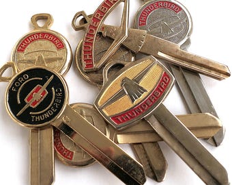 May include: A collection of six vintage gold-toned keys with red and black accents. The keys feature the words "Thunderbird" and "Ford" on the key tops.