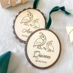 May include: Two wooden ornaments with a quail illustration and the names "Ava" and "Quinn" engraved. The ornaments have a scalloped edge and a dark brown border. Each ornament has a green velvet ribbon for hanging.
