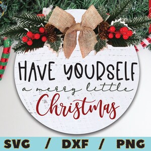 Puede incluir: Un letrero redondo de madera blanca con un fondo rojo desgastado y el texto "HAVE YOURSELF a merry little Christmas". El letrero est&aacute; decorado con un lazo marr&oacute;n, ramas de pino y bayas rojas.