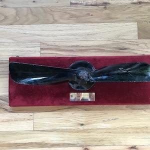 May include: A black propeller mounted on a red velvet base with a brass plaque. The propeller is mounted on a wooden base.