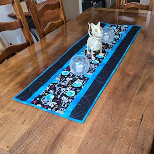 May include: A black and blue table runner with a skeleton cat design and teal accents. The runner is on a wooden table with two glass candle holders.