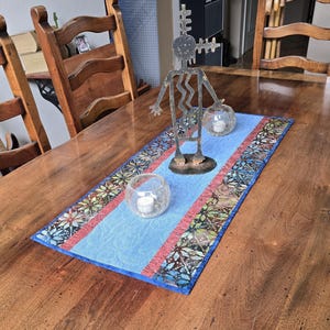 May include: A blue and red table runner with a geometric pattern. The runner is on a wooden table with a metal sculpture of a Kokopelli figure in the center. Two glass candle holders are on the table runner.