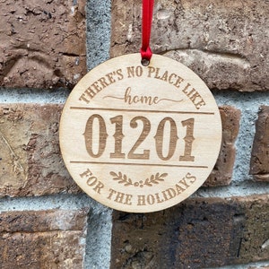 May include: A round wooden ornament with the text "There's no place like home" and the year "01201" in a decorative font. The ornament also has the text "For the Holidays" and a small wreath design.