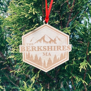 May include: Wooden Christmas ornament with a red ribbon. The ornament is shaped like a hexagon and features a mountain range with the text "THE BERKSHIRES MA" in a brown font.