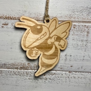 May include: A wooden ornament shaped like a cartoon bee with a yellow and black striped body. The bee is in a flying pose with its wings outstretched. The ornament has a string attached to the top for hanging.