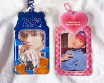 Puede incluir: Dos portafotos de acrílico transparente. Uno es azul con un diseño de oso de peluche y el texto "Teddy Bear". El otro es rosa con una parte superior en forma de corazón y el texto "i think i'm in love with you" y "my designer".