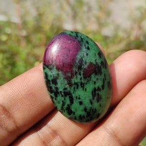 Può includere: Una pietra preziosa ovale di colore verde e rosa con macchie nere. La pietra preziosa è liscia e lucidata.