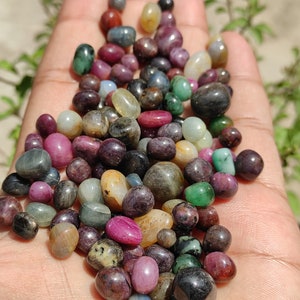 May include: A collection of tumbled gemstones in various colors, including red, pink, green, blue, and brown. The stones are smooth and polished, and they are arranged in a pile on a hand.