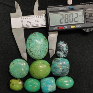 May include: A collection of eight turquoise cabochons, ranging in size and color, with the largest measuring 28.02 mm. The stones are displayed on a black surface with a digital caliper for size reference.