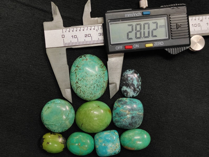 May include: A collection of eight turquoise cabochons, ranging in size and color, with the largest measuring 28.02 mm. The stones are displayed on a black surface with a digital caliper for size reference.