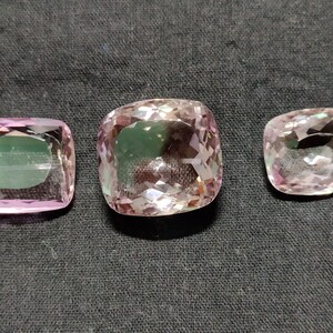 May include: Three pink amethyst gemstones of different sizes, cut in a cushion shape. The gemstones are faceted and have a translucent appearance.
