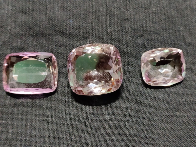 May include: Three pink amethyst gemstones of different sizes, cut in a cushion shape. The gemstones are faceted and have a translucent appearance.