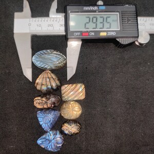 May include: A selection of carved labradorite gemstones in diverse shapes, such as oval, shell, and triangular. A digital caliper displays 29.35 mm. The gemstones exhibit iridescent blue and gold colours. The image shows a variety of carved designs.