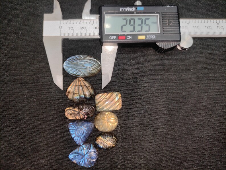May include: A selection of carved labradorite gemstones in diverse shapes, such as oval, shell, and triangular. A digital caliper displays 29.35 mm. The gemstones exhibit iridescent blue and gold colours. The image shows a variety of carved designs.