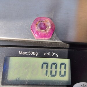 May include: A pink and white hexagonal gemstone with a star-shaped pattern inside. The gemstone is on a digital scale that reads 1.000.