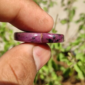May include: A close-up of a pink and black gemstone cabochon. The cabochon is round and has a smooth, polished surface. It is held in a person's hand.
