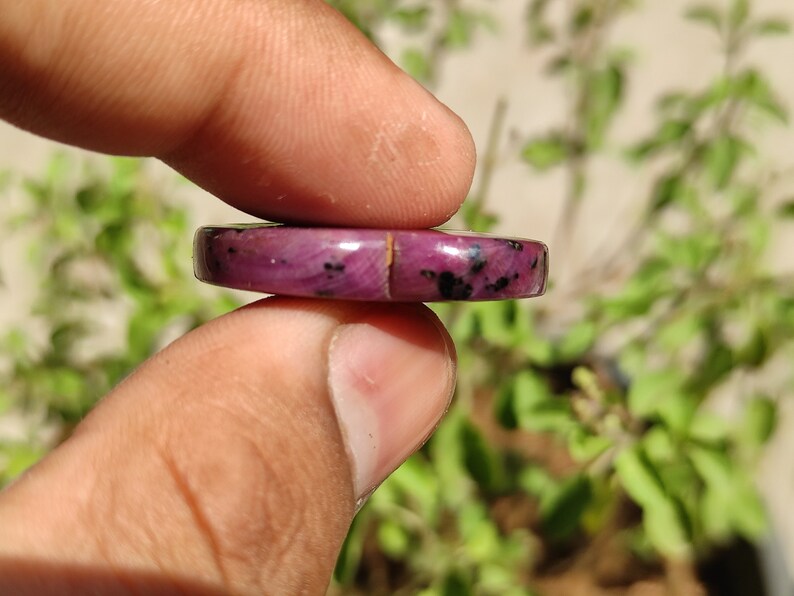 May include: A close-up of a pink and black gemstone cabochon. The cabochon is round and has a smooth, polished surface. It is held in a person's hand.